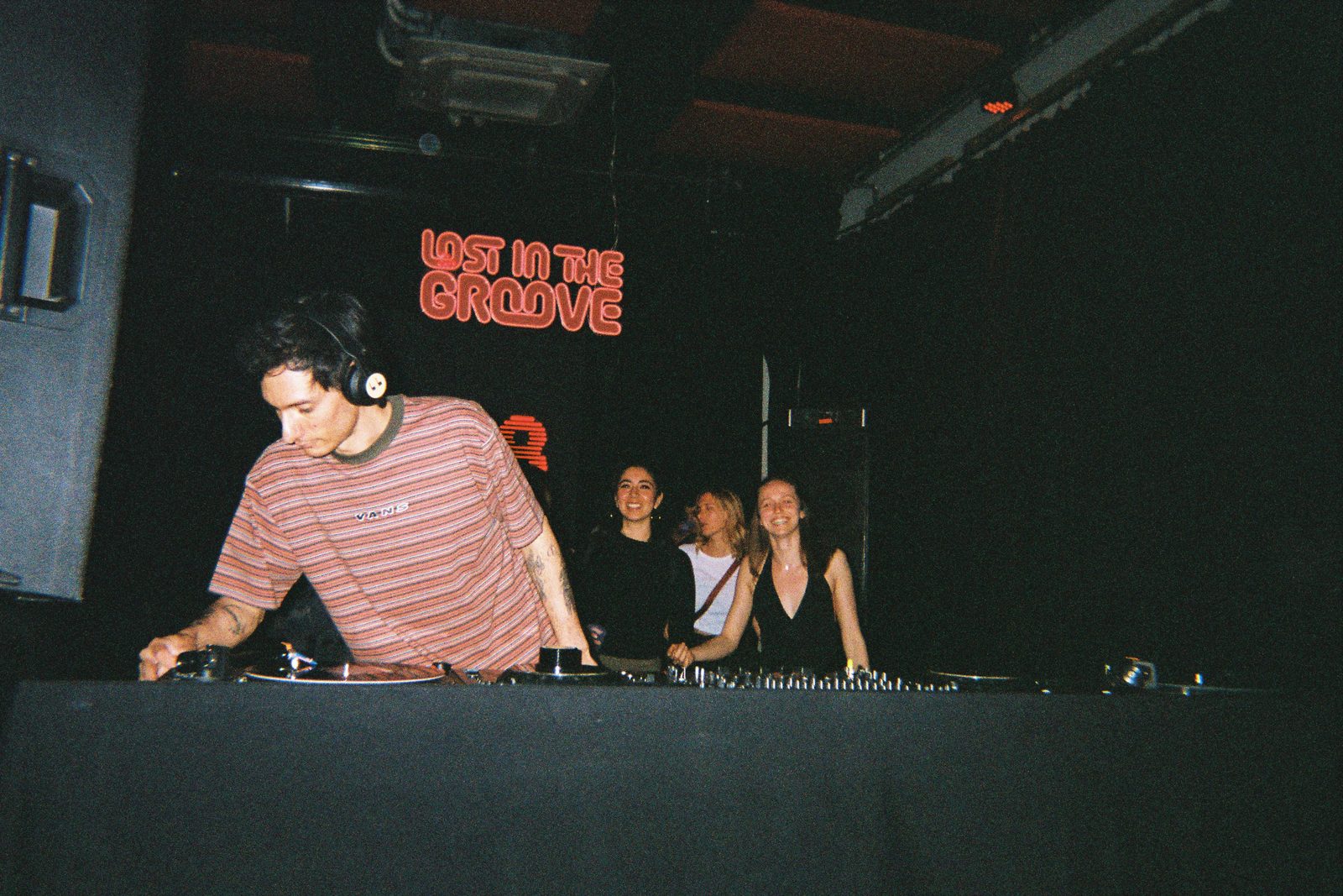 Fritz mezclando, tres amigas detrás, neón LITG